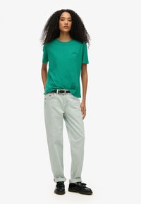 Groene t-shirt met ronde hals en een subtiel logo, gecombineerd met lichtblauwe, rechte jeans. Zwarte schoenen met een textuurafwerking maken de look compleet.
