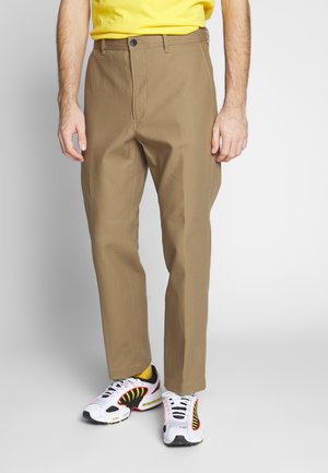 Mand iført beige bukser med lige ben, gule sokker, hvide og sorte sneakers og en gul skjorte, stående mod en neutral baggrund.