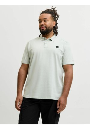 Mężczyzna z dredami i brodą, ubrany w jasnoszary polo z krótkim rękawem i czarne spodnie, stojący na jednolitym tle.