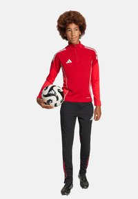 Rotes, langärmeliges Sportoberteil mit Reißverschluss und weißen Akzenten, kombiniert mit schwarzen Hosen mit roten Streifen. Hält einen schwarz-weißen Fußball.