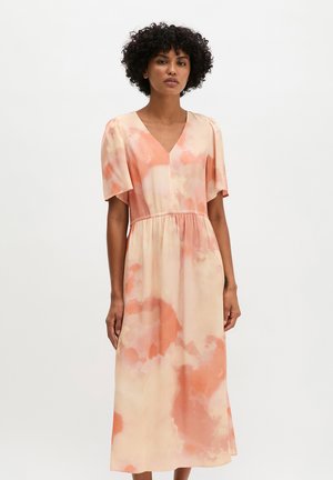 Frau mit lockigem Haar, die ein pfirsich- und cremefarbenes V-Ausschnitt-Kleid mit kurzen Ärmeln im Batik-Stil trägt, steht vor einem schlichten weißen Hintergrund.