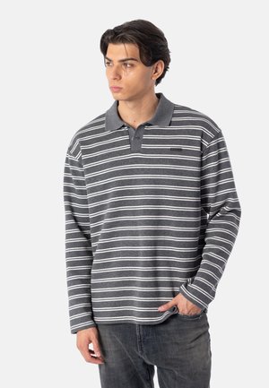 Jonge man met donker haar die een grijs-wit horizontaal gestreepte longsleeve poloshirt en donkere jeans draagt en opzij kijkt.