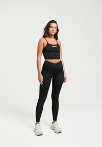 Zwarte sportieve crop top met dunne bandjes, gecombineerd met hoge zwarte leggings met een kruis ontwerp aan de voorkant, gedragen met witte sneakers.