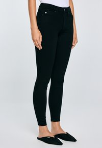 Cilvēks, kurš valkā melnas pieguļošas bikses, baltu augšu un melnas norādītas slip-on kurpes, stāv ar rokām brīvi gar sāniem.