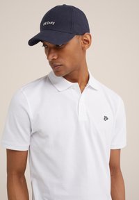 Camisa polo blanca de manga corta con un acabado texturizado, que presenta un pequeño logotipo negro en el lado izquierdo del pecho; combinada con una gorra azul marino.