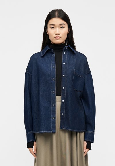 Femme aux longs cheveux foncés portant une veste en denim foncé sur un col roulé noir et une jupe beige, debout devant un fond blanc uni.