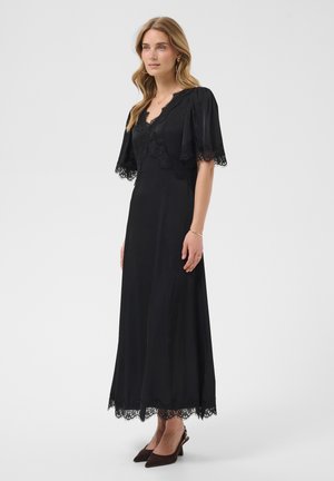 Mujer de pie con un vestido negro hasta los tobillos con ribete de encaje, mangas cortas con vuelo y tacones oscuros, sobre un fondo liso y claro.