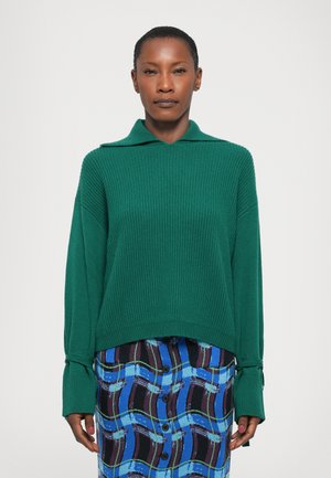 Maglione verde a coste con colletto e maniche lunghe. Abbinato a una gonna blu, viola e nera con un motivo a fantasie e dettagli con bottoni.