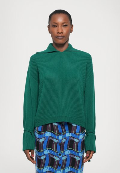Pull en tricot côtelé vert avec un col et des manches longues. Associé à une jupe à motifs bleus, violets et noirs avec des détails de boutons.