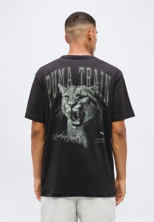 Mężczyzna z krótkimi włosami, odwrócony tyłem, nosi czarną koszulkę z ryczącym pumą i tekstem "Puma Train" na plecach.