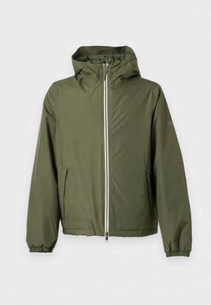 Jeune homme aux cheveux tressés portant une veste à capuche verte, une chemise couleur rouille, un pantalon cargo beige et des chaussures marron, debout devant un fond uni.