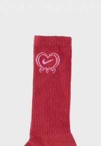 Raudonos juostos kojinė, ant kurios yra rausvos širdies ir „Swoosh“ logotipo dizainas. Audinys atrodo minkštas ir lygios tekstūros.