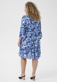 Blaue gemusterte Kleid mit 3/4-Ärmeln, gestuften Rüschen und weichem, leichtem Stoff; enthält ein florales Design in verschiedenen Blautönen.