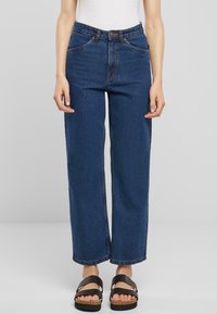 Jeans de pierna ancha azul oscuro con una cintura alta, bolsillos frontales y costuras en contraste, combinados con sandalias negras de tiras y una blusa ajustada blanca.
