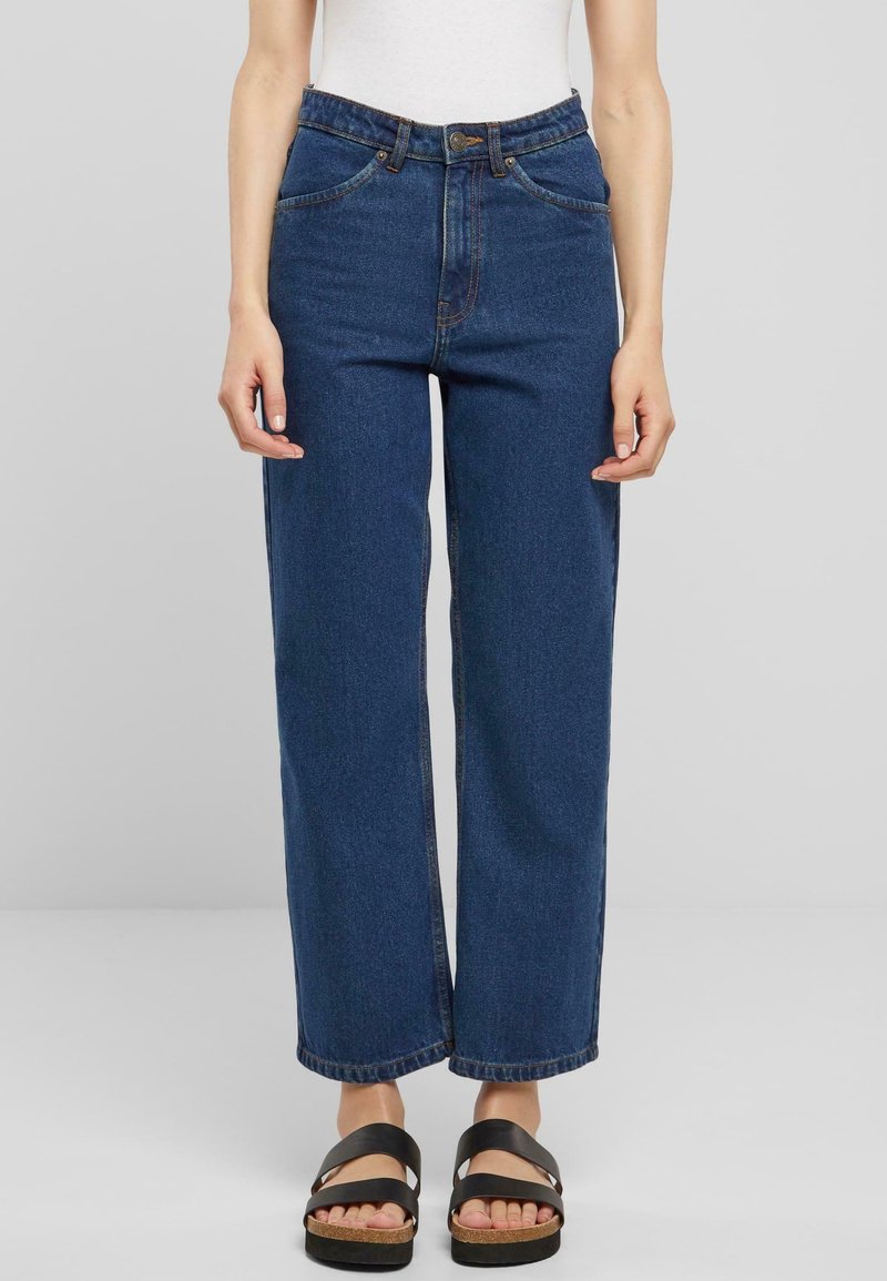 Jeans de pierna ancha azul oscuro con una cintura alta, bolsillos frontales y costuras en contraste, combinados con sandalias negras de tiras y una blusa ajustada blanca.