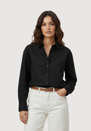 Vrouw met lang bruin haar, draag een zwart overhemd met knopen, gestopt in een witte broek met een bruine riem, staand met één hand in de zak.