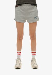 Pantalones cortos de algodón gris con una cinturilla elástica que presenta bordado en azul marino "SUPERDRY NYC" a la izquierda. Combinados con calcetines blancos y zapatillas.