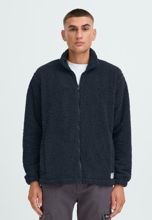 Schwarze, Fleece-Zip-Jacke mit hohem Kragen, aus strukturierter Stoff gefertigt und mit langen Ärmeln; getragen über einem weißen Hemd, mit einem Logosticker am unteren Ende.