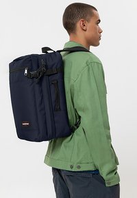 Mochila de color azul marino con forma rectangular, fabricada en un tejido duradero. Cuenta con un bolsillo frontal con cremallera y correas ajustables negras.