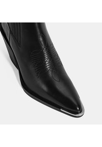 Bottines en cuir noir à bout pointu, avec des motifs en relief et un accent métallique sur le capuchon.