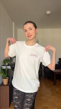 Jonge vrouw in een wit Diesel T-shirt en een zwarte kanten rok staat binnen, houdt de kraag van het shirt bij de schouders vast, met planten en meubels op de achtergrond.