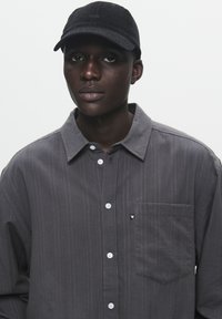 Gray collared shirt with vertical stripes, featuring a chest pocket and white buttons. Black cap with logo on front. Smooth texture.