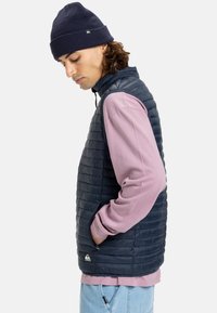 Junger Mann in einer marineblauen Steppweste, rosa Langarmshirt und marineblauer Mütze, der nach unten schaut, mit Händen in den Taschen der Weste vor weißem Hintergrund.