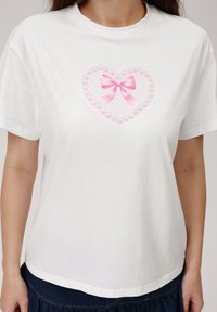 Camiseta de algodón blanca con un diseño de corazón hecho de perlas sintéticas y un lazo rosa en el centro. Mangas cortas y un ajuste relajado.