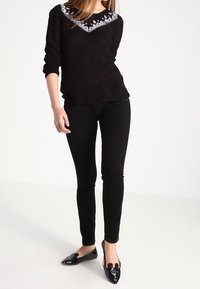 Mujer vestida con una blusa negra de manga larga con bordados florales blancos, pantalones ajustados negros y mocasines negros de punta, de pie contra un fondo blanco.