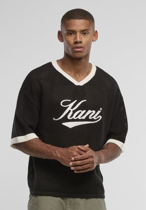 Tânăr purtând un tricou negru din plasă cu mâneci scurte, cu margine albă și logo "Kani", stând în fața unui fundal simplu gri deschis.