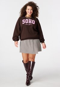 Bruine trui met roze tekst "SOHO New York", gecombineerd met een zwart-witte pied-de-poule rok en glanzende bruine kniehoge laarzen.