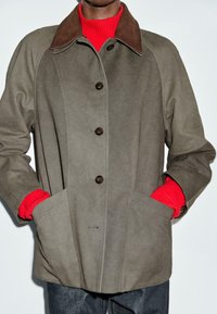 Persona che indossa un giaccone grigio con bottoni e colletto marrone su un maglione rosso brillante, mani in tasca e pantaloni scuri su sfondo bianco.