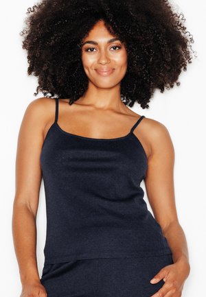 Vrouw met krullend haar glimlacht, gekleed in een aansluitend donkerblauw camisole top en bijpassende broek tegen een effen witte achtergrond.