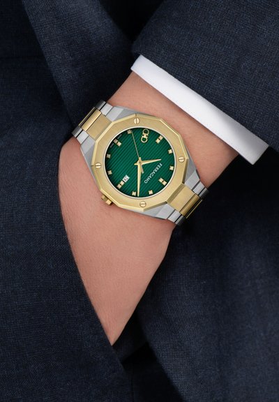 Zöld számlapú karóra arany és ezüst lünettával, gyémánt órakijelzőkkel, dátumkijelzéssel, bal csuklón viselve, elrejtve a tengerészkék öltönyzsebben.