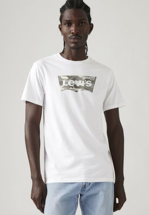 Jeune homme aux cheveux tressés portant un t-shirt blanc Levi's avec un logo camouflage et un jean bleu clair sur fond uni.