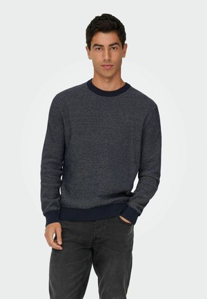 Jeune homme aux cheveux foncés portant un pull texturé bleu marine et un jean noir, debout avec une main dans la poche devant un fond uni.
