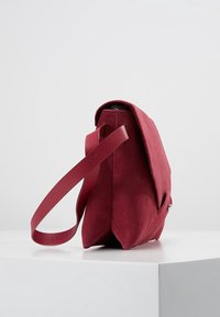 Červená semišová kabelka se strukturovaným designem, plochým dnem a jedním nastavitelným popruhem. Obsahuje klopový uzávěr a jemné kovové akcenty.