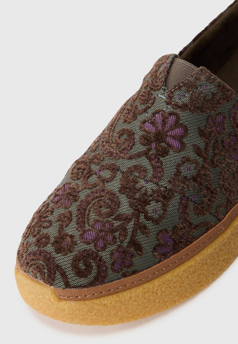 Zapato tipo slip-on con un upper de tela verde oscuro y bordados en flores marrones y moradas. Tiene una punta redondeada y una suela texturizada en color beige.