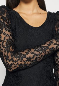 Schwarzes Spitzenoberteil mit durchsichtigen langen Ärmeln mit Blumenmustern, einem Rundhalsausschnitt und einem körperbetonten Schnitt, der die strukturierte Überlagerung bildet.