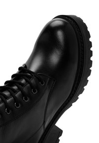 Bota negra de cuero hasta el tobillo con punta redondeada, superficie texturada y suela gruesa con tacos. Presenta ojales de metal y cordones negros.