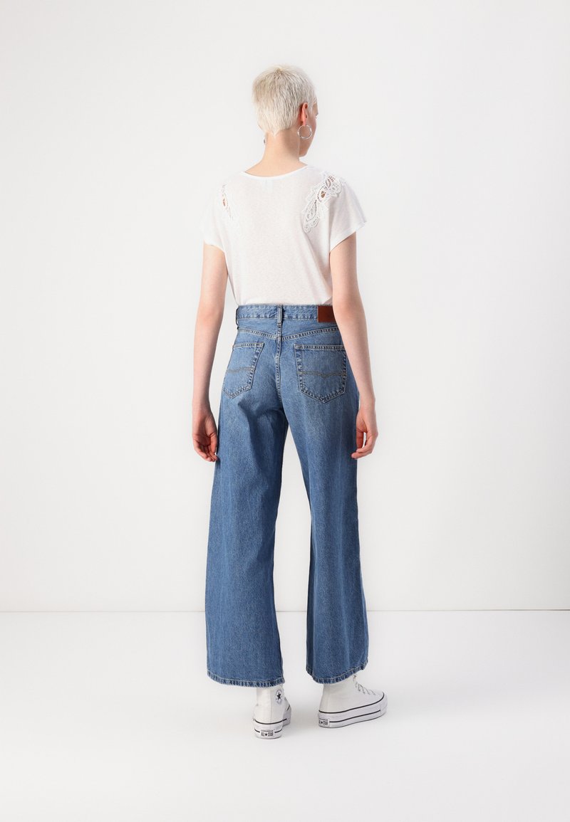 Jean denim à jambes larges en bleu clair, avec une taille haute, des poches arrière et un léger effet usé. Associé à un haut blanc, présentant des accents en dentelle.