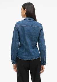 Temno modra denim jakna s prilegajočo zasnovo, vidnimi šivi in gumbastimi manšetami. Ima klasičen ovratnik in tapered obliko na zadnjem delu.