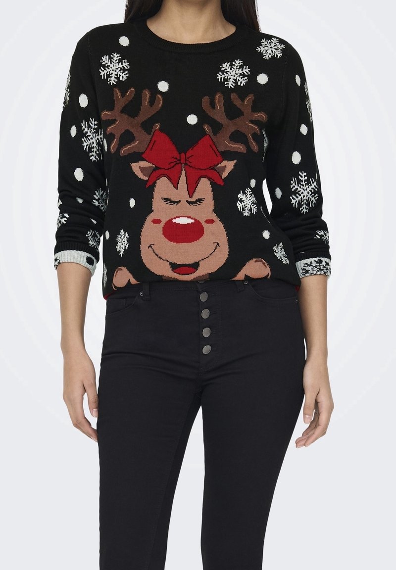 Femme portant un pull noir avec un visage de renne souriant, un nœud rouge et des motifs de flocons de neige, assorti à un pantalon noir boutonné.