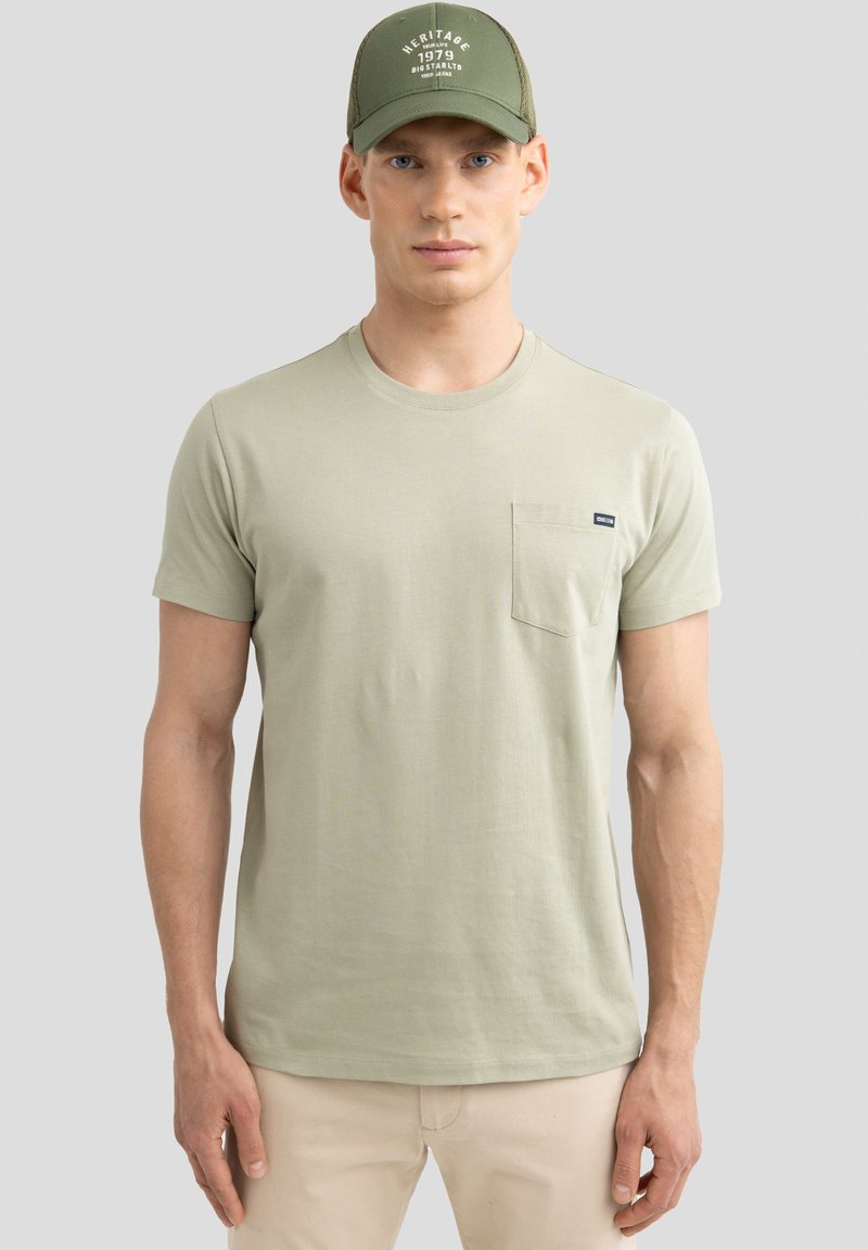 Šviesiai žalia trumpomis rankovėmis T-shirt marškiniai su apvaliu kaklu, krūtinės kišenė ir subtiliu tekstūru. Suderinta su žalia skrybėle su balta tekstu.