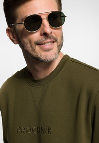 Zielony sweatshirt z haftowanym logo, z okrągłym dekoltem i teksturowanym materiałem. Okrągłe czarne okulary przeciwsłoneczne z metalowymi akcentami.