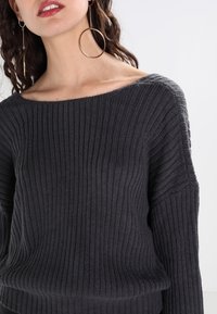 Pull en maille côtelée gris foncé avec un décolleté asymétrique, des manches longues et une coupe décontractée. Le tissu texturé présente des lignes verticales.