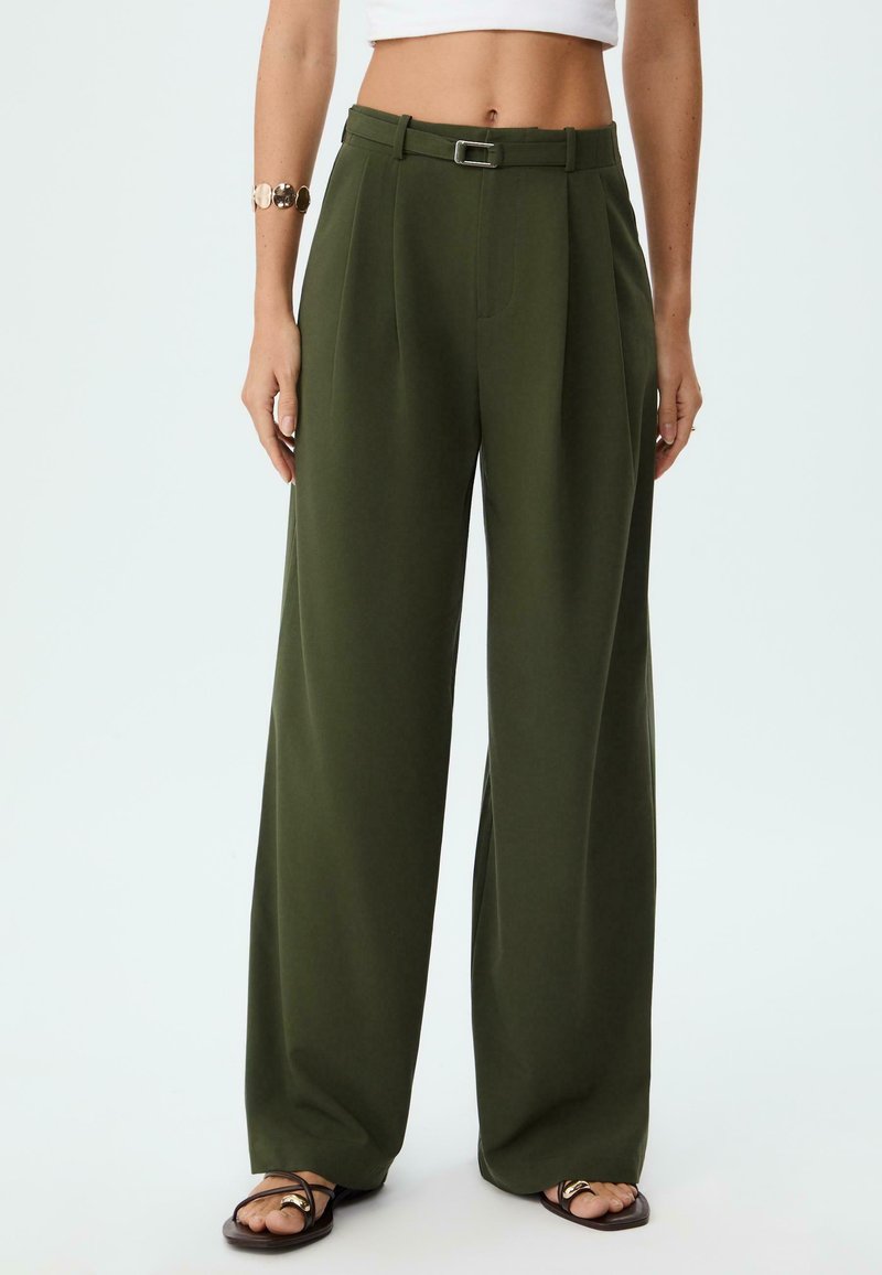 Pantaloni a gamba larga in tessuto verde scuro, con vita alta e cinturino, pieghe e una texture liscia, abbinati a sandali minimalisti.