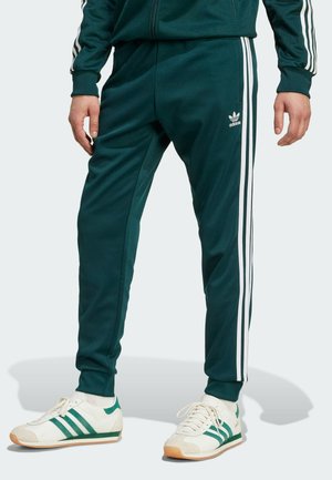Grønne atletiske bukser med hvide sidelister, lavet af glat stof. Bæres med hvide sneakers med grønne detaljer og gummisåler.