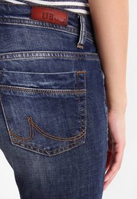 Jeansy z ciemnoniebieskiego denimu z pięcioma kieszeniami. Widoczne pomarańczowe przeszycia oraz skórzana naszywka LTB na pasku. Gładka faktura.