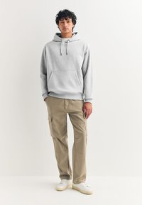 Sweat à capuche gris avec une poche kangourou, associé à un pantalon cargo beige avec des poches latérales et des baskets blanches sur un fond neutre.